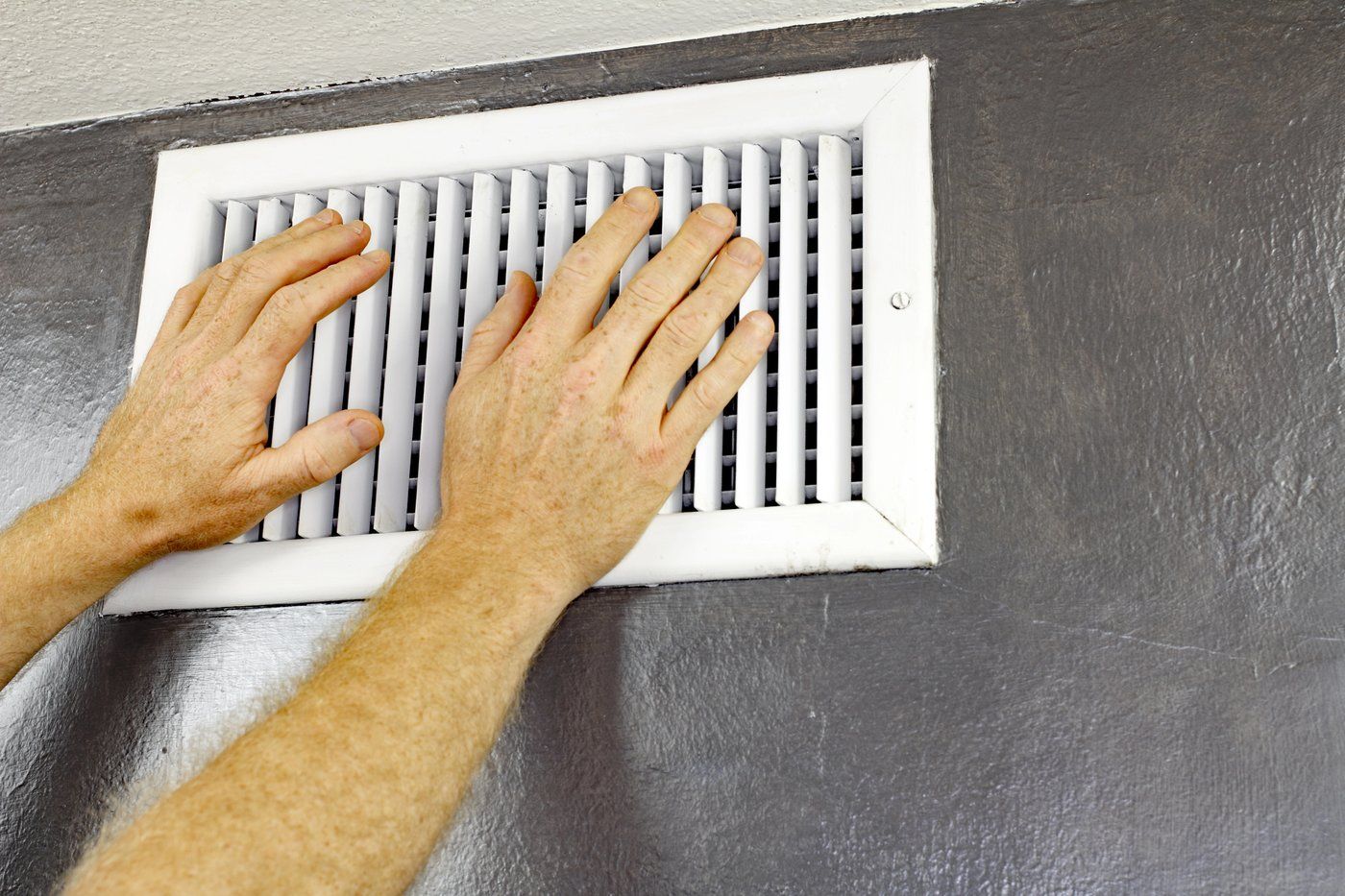 Two hands feeling for airflow at a white wall-mounted heating and cooling vent