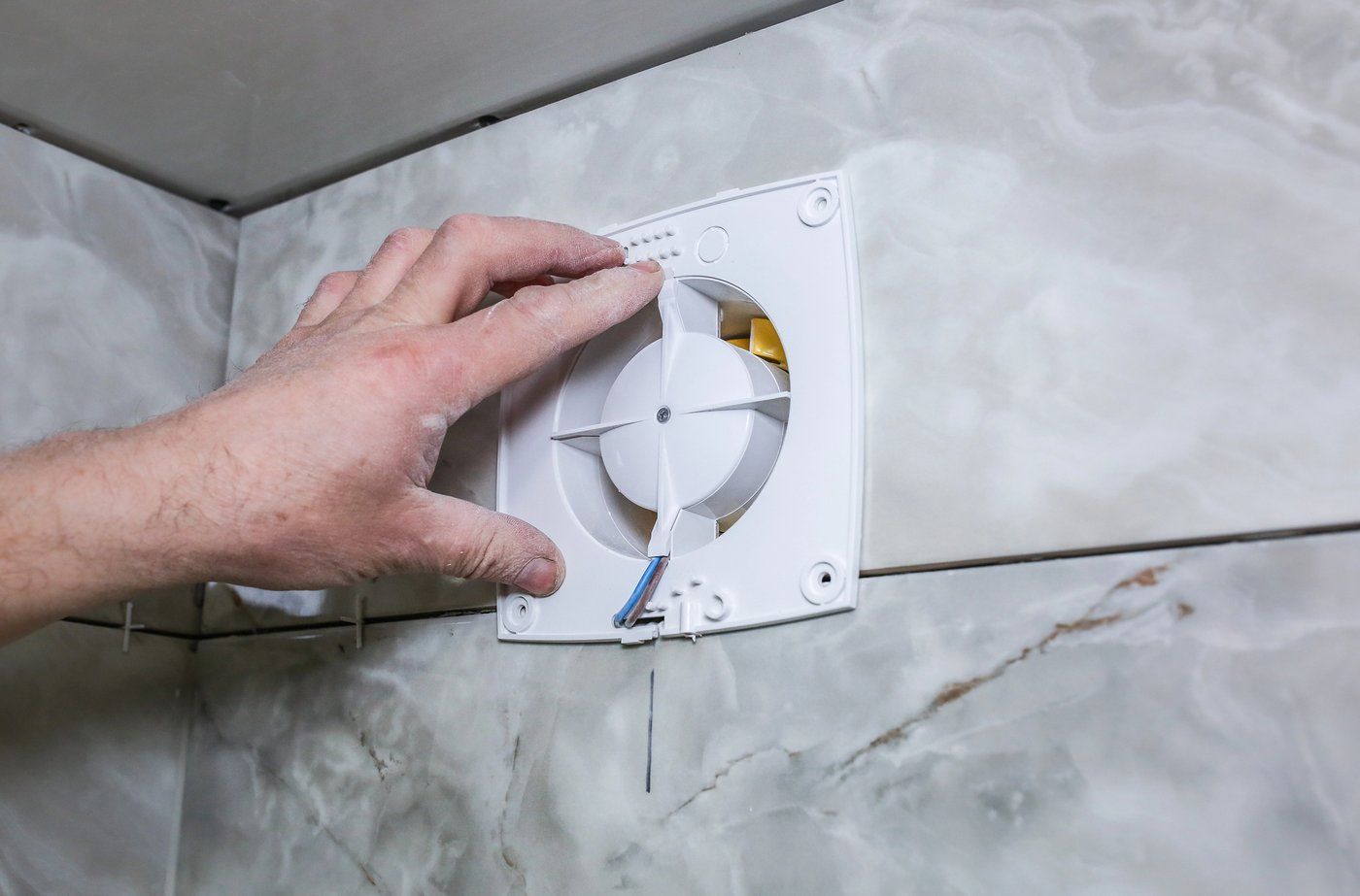 Hand installing a bathroom extractor fan on a marble-tiled wall
