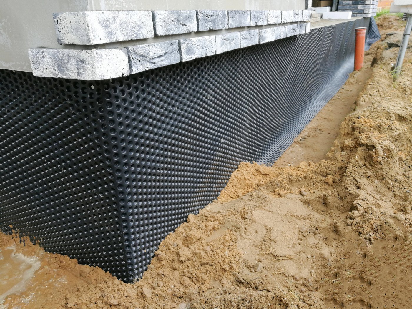 Black dimpled waterproofing membrane fixed to a foundation wall with an orange drainage pipe