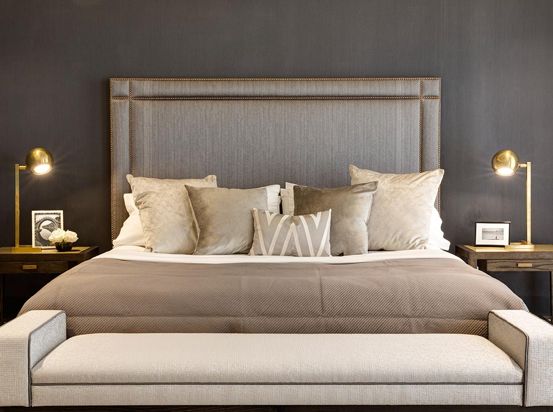 bedroom with large bed with bed bench, grey wall and bedside tables with gold lamps