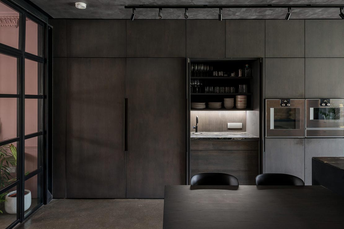 dark brown modern kitchen with wall ovens, integrated fridge and small dining area